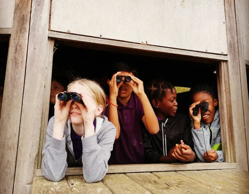 Four smiling children at a bird hide, chatting and looking through binoculars as they enjoy birdwatching together.